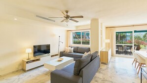 A modern living room with a flat-screen TV, grey sofas, a coffee table, and a ceiling fan. - Lunamar Garden first line beach Real de Zaragoza (Marbella)