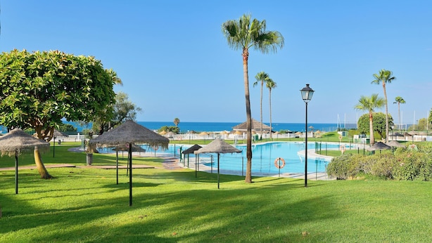 A pool area with palm trees, a clear sky, and a view of the ocean. - Lunamar Garden first line beach Real de Zaragoza (Marbella)