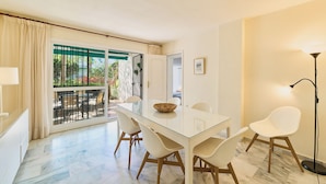 A modern dining area with a glass table, white chairs, and a view through a sliding glass door to an outdoor patio. - Lunamar Garden first line beach Real de Zaragoza (Marbella)