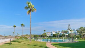 A pool area with a clear sky, palm trees, and a building with balconies in the background. - Lunamar Garden first line beach Real de Zaragoza (Marbella)
