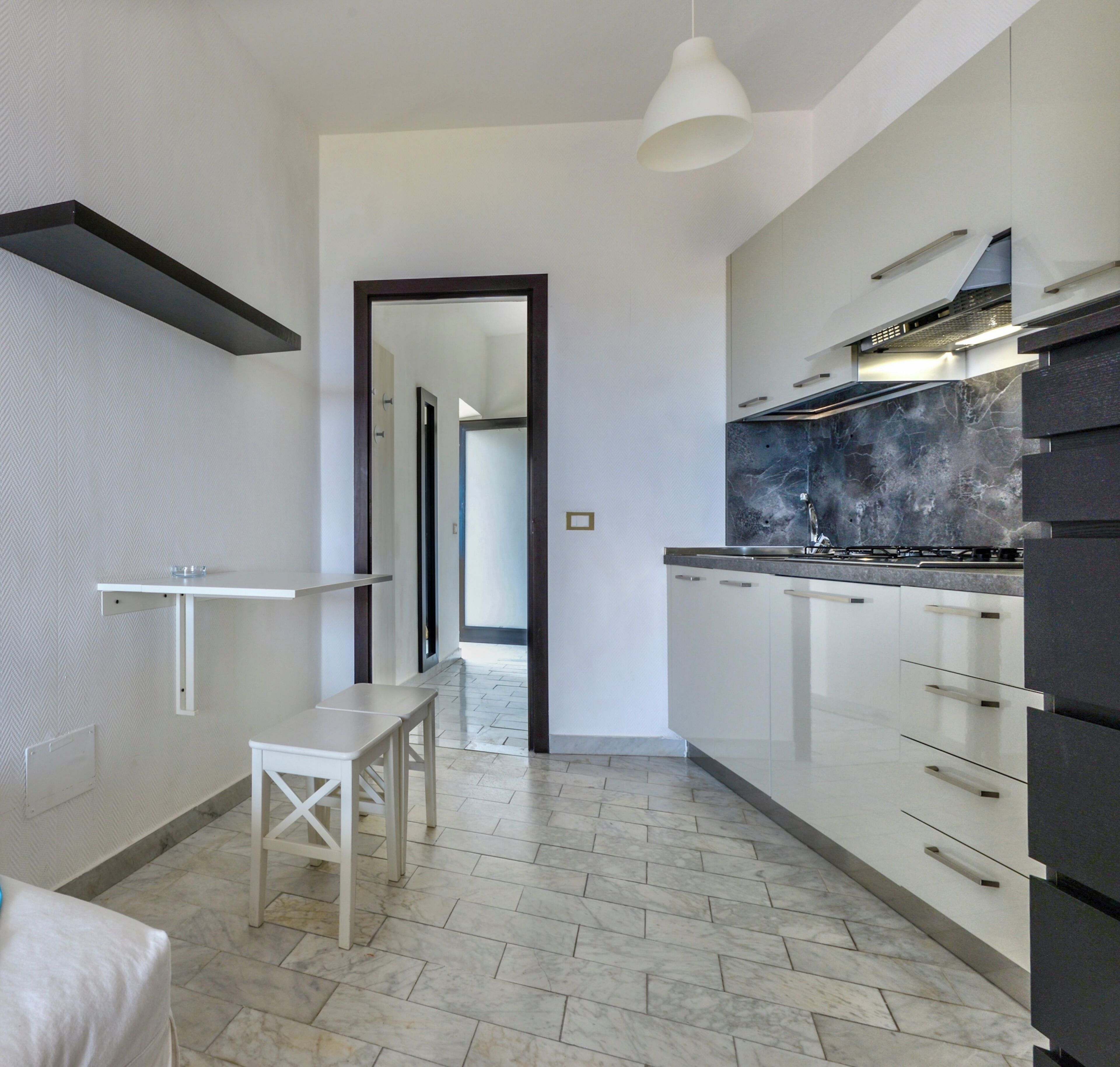A modern kitchen with white cabinets, a marble countertop, and a black backsplash. There is a white bench and a hanging light fixture.