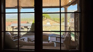 A room with a large window overlooking a patio area with outdoor furniture and a view of a building with a red roof. - LOS RELOJES HOUSE (La Adrada)