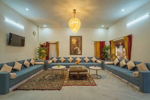 A living room with a blue sofa set, a chandelier, a flat-screen TV, and a large painting on the wall. - Hotel Narpat Garh Palace Jaisalmer (Jaisalmer)
