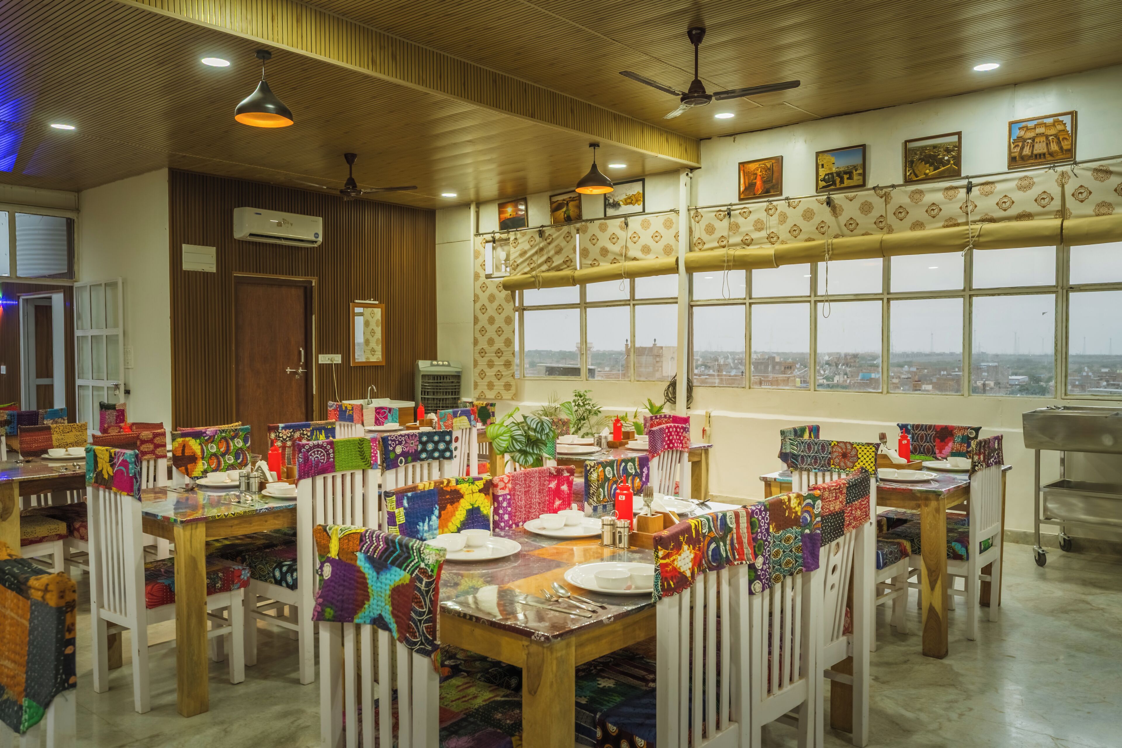 Un espace repas avec des tables dressées pour les repas, des chaises recouvertes de tissus aux couleurs vives et une vue dégagée sur l’extérieur grâce à de grandes fenêtres.
