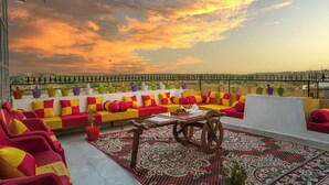 A rooftop terrace with a sofa set, a table with a teapot, and a view of the cityscape during sunset. - Hotel Narpat Garh Palace Jaisalmer (Jaisalmer)