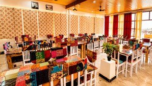 A dining area with tables covered in colorful tablecloths, white chairs, and a large potted plant. - Hotel Narpat Garh Palace Jaisalmer (Jaisalmer)