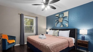 Quarto com cama, mesa de cabeceira, cadeira, ventoinha no teto e janela com cortinas.