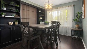 Uma área de refeições com mesa e cadeiras de madeira, cercada por armários de madeira escura com prateleiras que exibem plantas e objetos decorativos.