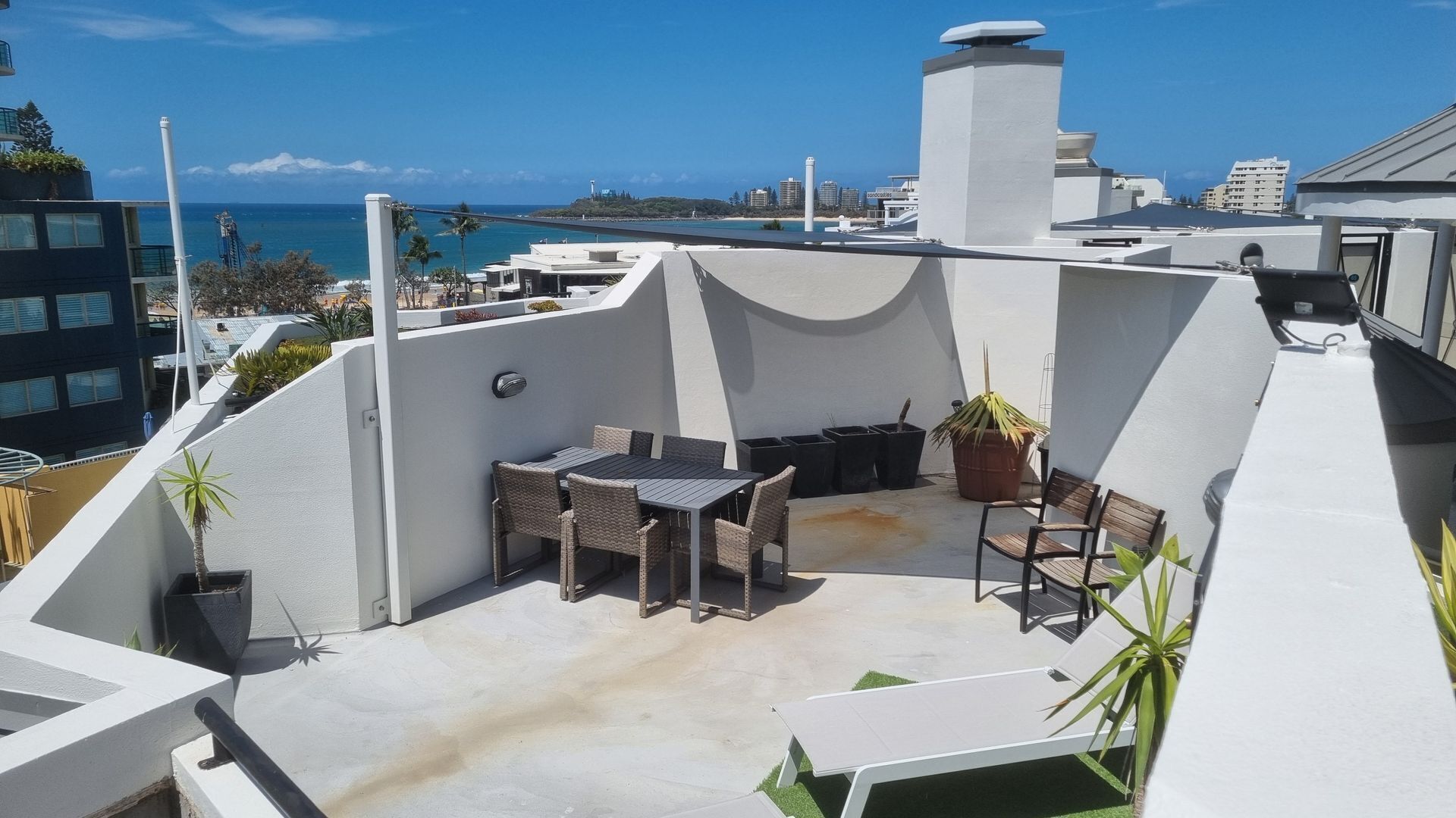 A rooftop terrace with a dining area, potted plants, and a view of the ocean and city skyline.