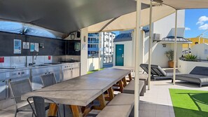 An outdoor kitchen area with a long table and chairs, a grill, and a view of a building with a blue door. - 62 at the Caribbean Resort (mooloolaba)