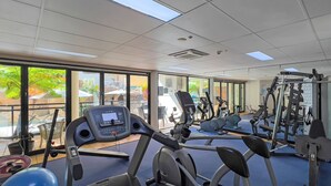 A gym with cardio machines, weight equipment, and large windows offering views of outdoor scenery. - 62 at the Caribbean Resort (mooloolaba)