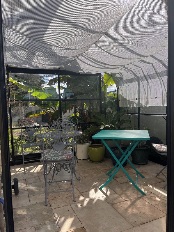 Área de varanda coberta com mesa e cadeiras de metal, plantas em vasos e vista para o verde lá fora.