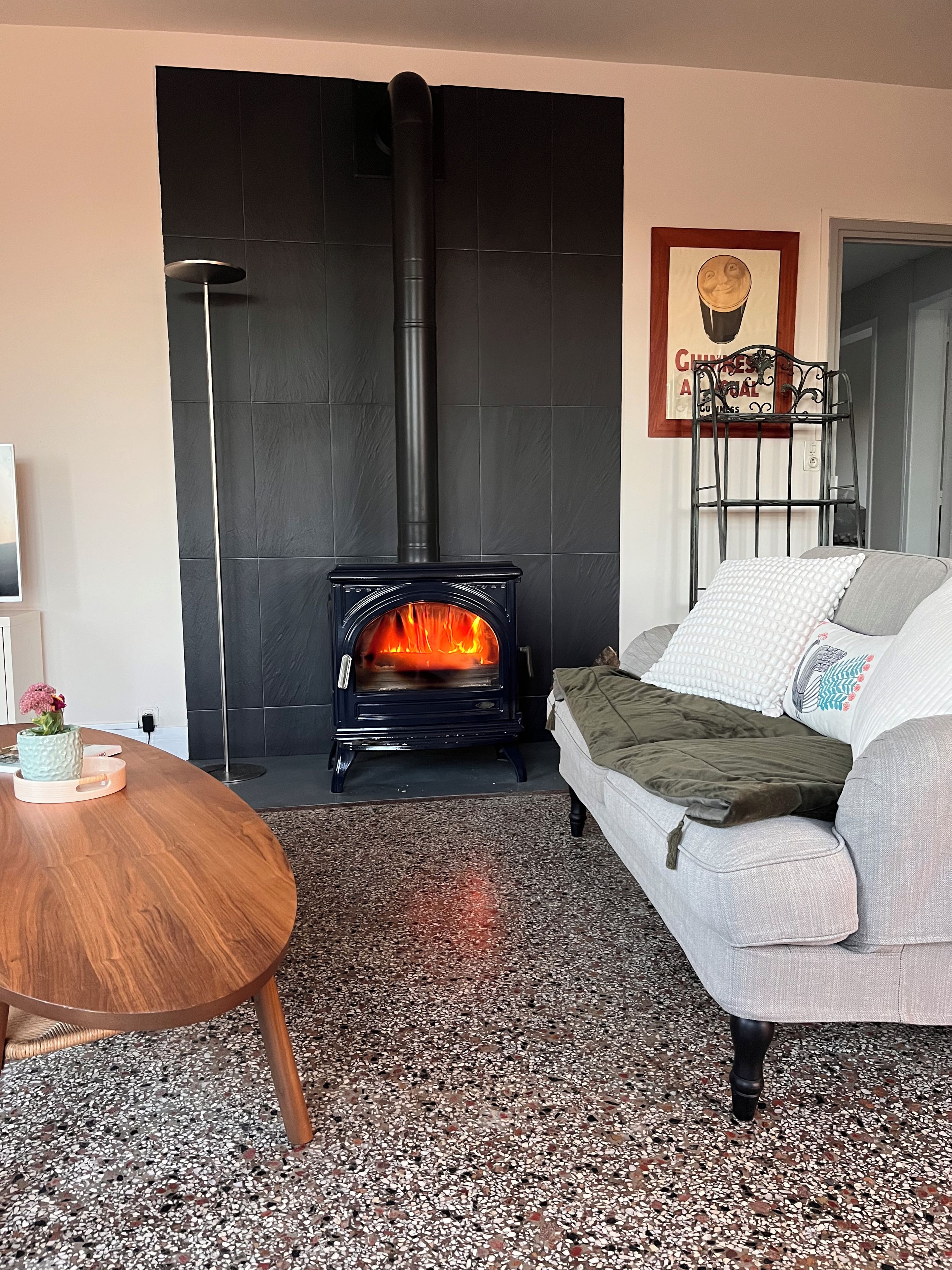 Ein Wohnzimmer mit schwarzer Fliesenwand, einer Couch, einem runden Holztisch und einem Kamin mit sichtbaren Flammen.