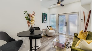 A modern living room with a round table, a sofa, and a chair. There is a ceiling fan, a painting on the wall, and a window with blinds. - Tiny Home Atlanta Retreat (Union City)