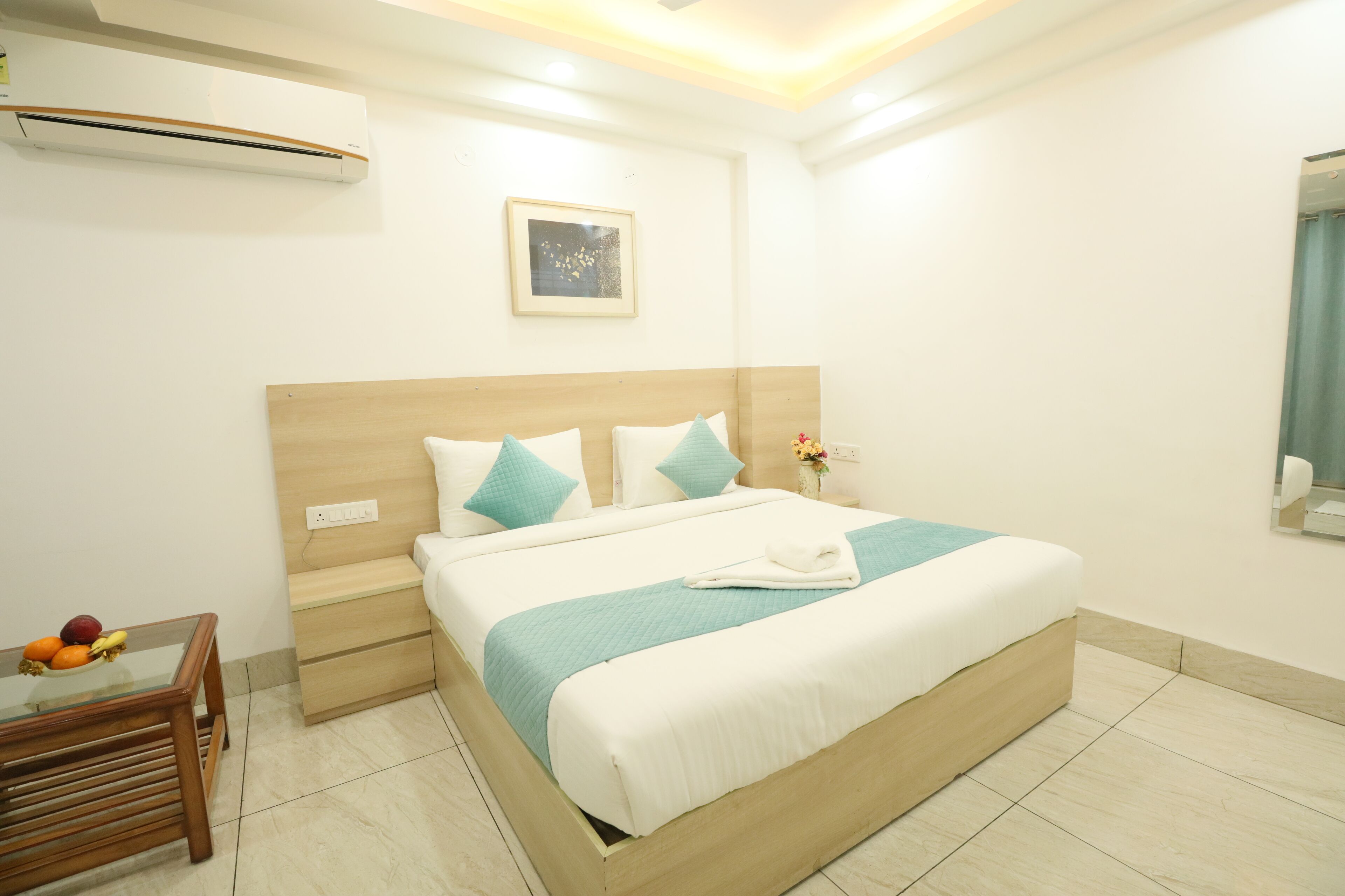 A hotel room with a large bed, a bedside table with fruit, a wall-mounted air conditioner, and a framed picture above the bed.