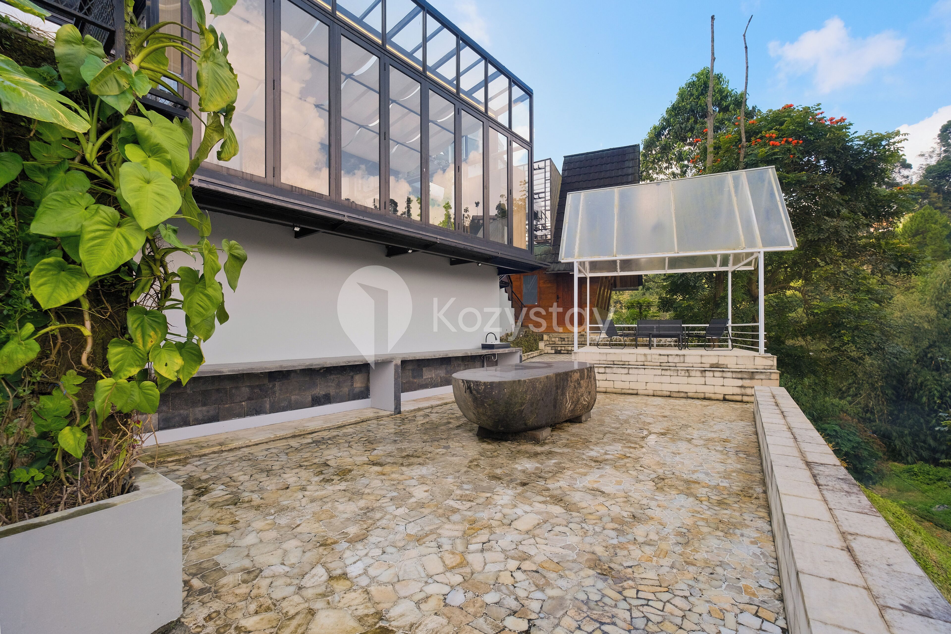A modern outdoor patio with a stone floor, a large planter with green plants, and a seating area with a bench and a table.