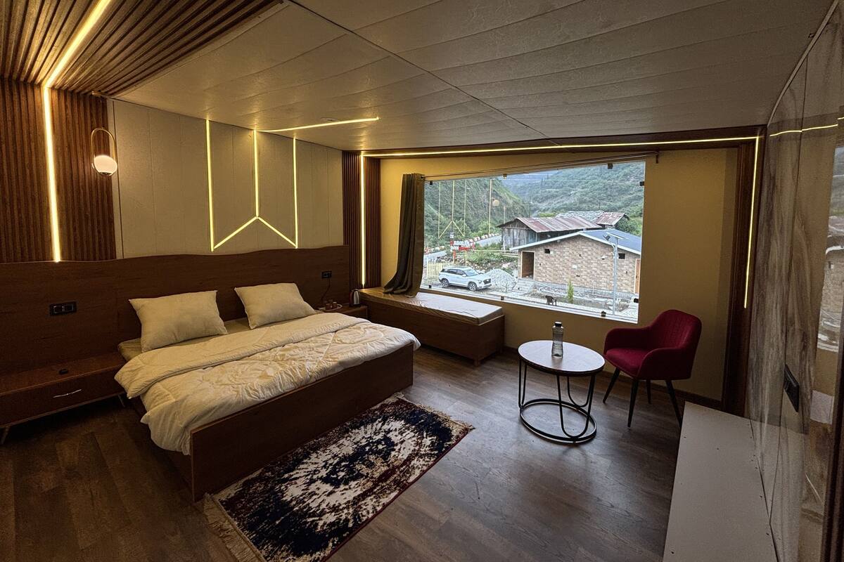 A modern bedroom with a large bed, a small table, a chair, and a view of a village through a window.