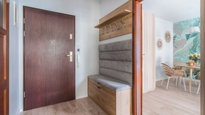 Un intérieur moderne avec une porte en bois, un banc avec espace de rangement, un coin repas avec des chaises en osier et un mur orné d’un motif de feuilles.