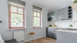 Une cuisine moderne avec des meubles gris, un four à micro-ondes et un évier. On y trouve deux fenêtres équipées de stores, un radiateur et une petite table sur laquelle sont disposés des fruits.