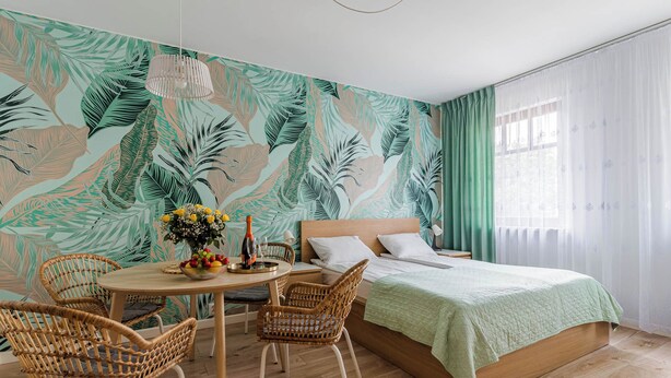 Une chambre à coucher avec un lit, une table ronde entourée de chaises, un vase de fleurs et un mur décoré d’un motif de feuilles tropicales.