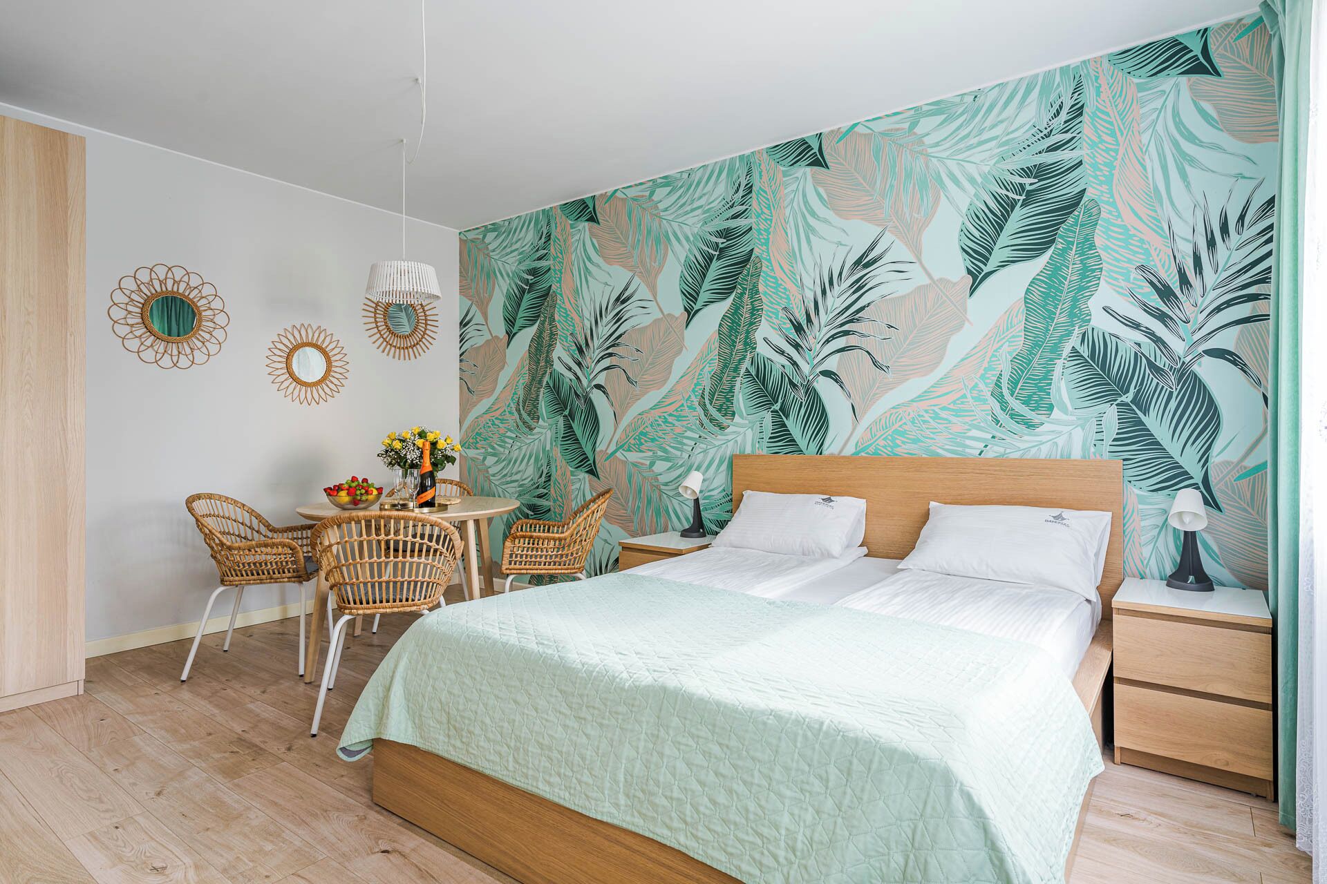 Une chambre avec un grand lit, une petite table avec des chaises et un mur orné d’un motif de feuilles tropicales.