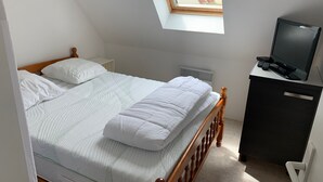 A bedroom with a bed, a desk with a computer, and a window with a view of a red-roofed building. - Charming Cottage - Near Beach and Shops (Fort-Mahon-Plage)