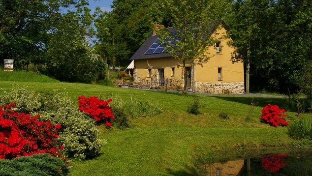 A house with a stone foundation and a yellow exterior, surrounded by lush greenery and red flowers. - Detached house with terrace, pellet stove and peaceful garden. (Canisy)