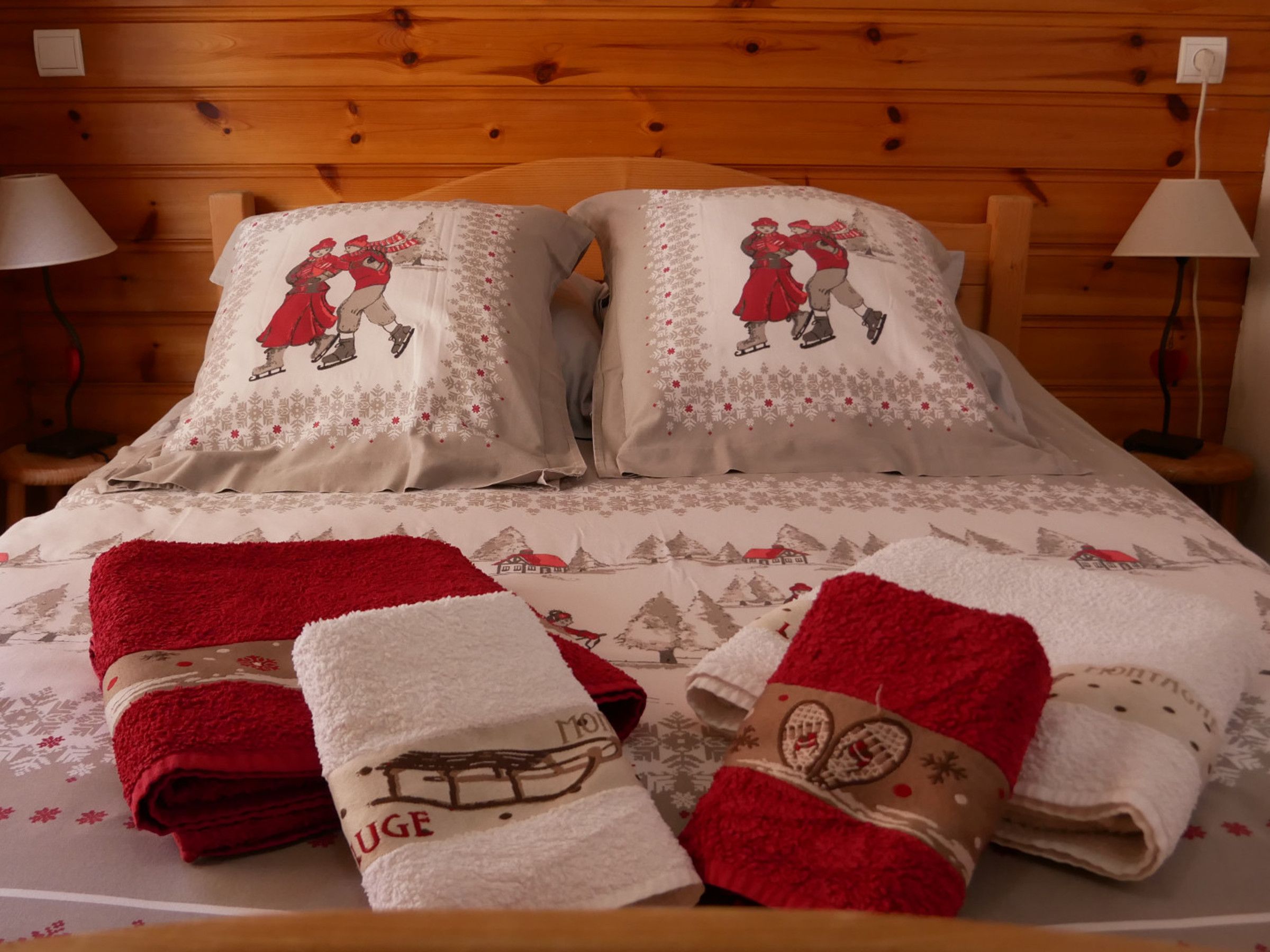 A bed with two pillows featuring a winter scene, a red blanket, and two towels with a mountain scene.