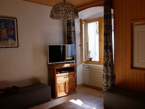 A living room with a television on a wooden cabinet, a sofa, a window with curtains, and a wall-mounted lamp. - Spacious apartment in Samoëns, 3 bedrooms, WiFi, 10 people (Samoëns)