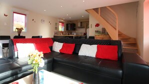 Een woonkamer met een zwarte leren bank, een eettafel met stoelen, een keuken op de achtergrond en een houten trap.