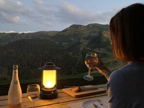 A person seated at a wooden table with a bottle, a glass, and a lit lantern, overlooking a mountainous landscape.