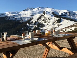 Une table de pique-nique en bois, dressée avec des aliments pour le petit-déjeuner, comme des œufs, du pain et des boissons, et offrant une vue sur une montagne enneigée.