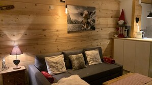 Ein gemütliches Zimmer mit einer Holzwand, einem grauen Sofa mit dekorativen Kissen, einem kleinen Nachttisch mit Lampe und einer Küchenzeile mit herzförmiger Dekoration.