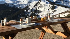 Ein Holztisch mit Frühstücksartikeln wie Eiern, Brot und Getränken, mit Blick auf einen schneebedeckten Berg.
