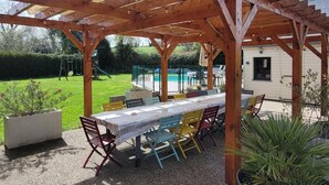 A covered patio area with a table and chairs, a pool in the background, and a house. - Large family home with private pool, pets allowed (Isigny-le-Buat)