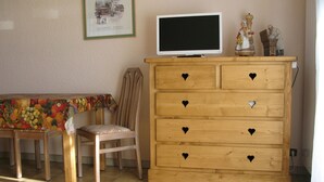 A wooden dresser with heart-shaped handles, a television on top, and a bench with a fruit-patterned tablecloth.
