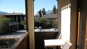 A balcony with a wooden bench and table, overlooking a parking area and buildings with a mountain in the background.