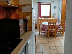 A kitchen with a stove, a dining table with chairs, a window with curtains, and a wooden cabinet. - Cozy 2-room apartment with Wifi in Samoëns (Samoëns)