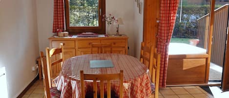 A dining area with a round table covered by a checkered tablecloth, wooden chairs, a wooden cabinet, and a window with a view of greenery.