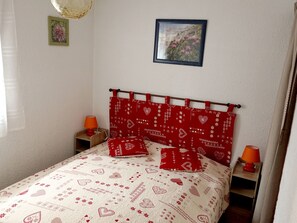 Un dormitorio con cama, dos mesitas de noche, un tocador, una ventana con cortinas y una pared con dos cuadros enmarcados.