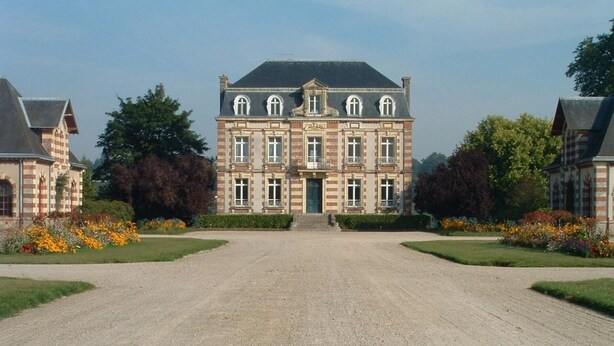 A large, symmetrical building with a central entrance, flanked by smaller structures, surrounded by well-maintained gardens and a gravel driveway.