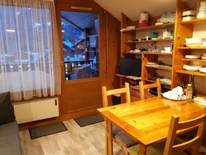 A wooden dining table with chairs, a television, and a kitchen area with shelves and a window.