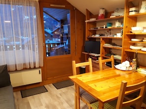 A wooden dining table with chairs, a television, and a kitchen area with shelves and a window. - Cozy studio for 2 in Samoëns (Samoëns)