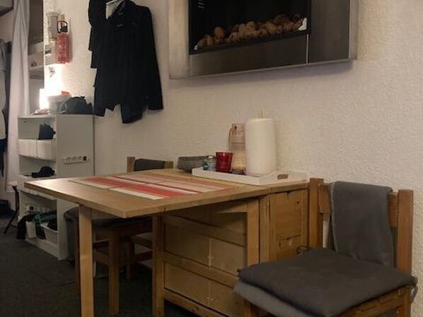 A small room with a wooden table, a chair, a shelf with various items, and a wall-mounted shelf with a few objects.