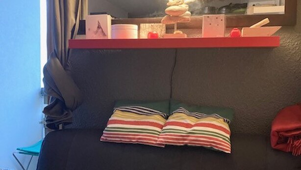 Une chambre avec une tête de lit noire, des coussins à rayures, une étagère rouge sur laquelle sont disposés divers objets, et une fenêtre avec des rideaux.