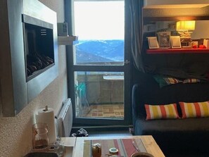 A hotel room with a bed, a small table with a lamp, a window with a view, and a shelf with items.