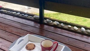 Une terrasse en bois avec une rambarde, une assiette avec un biscuit et une tasse, et une vue sur un espace verdoyant avec des arbres.