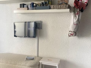 A room with a white textured wall, a small white side table, a beige sofa, a wall-mounted shelf with various items, and a hanging bag with a red and white pattern.