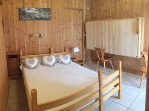 Quarto de madeira com uma cama, uma cadeira, uma escrivaninha e uma televisão presa à parede.