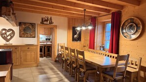 A wooden interior with a long dining table, chairs, and a kitchen area.